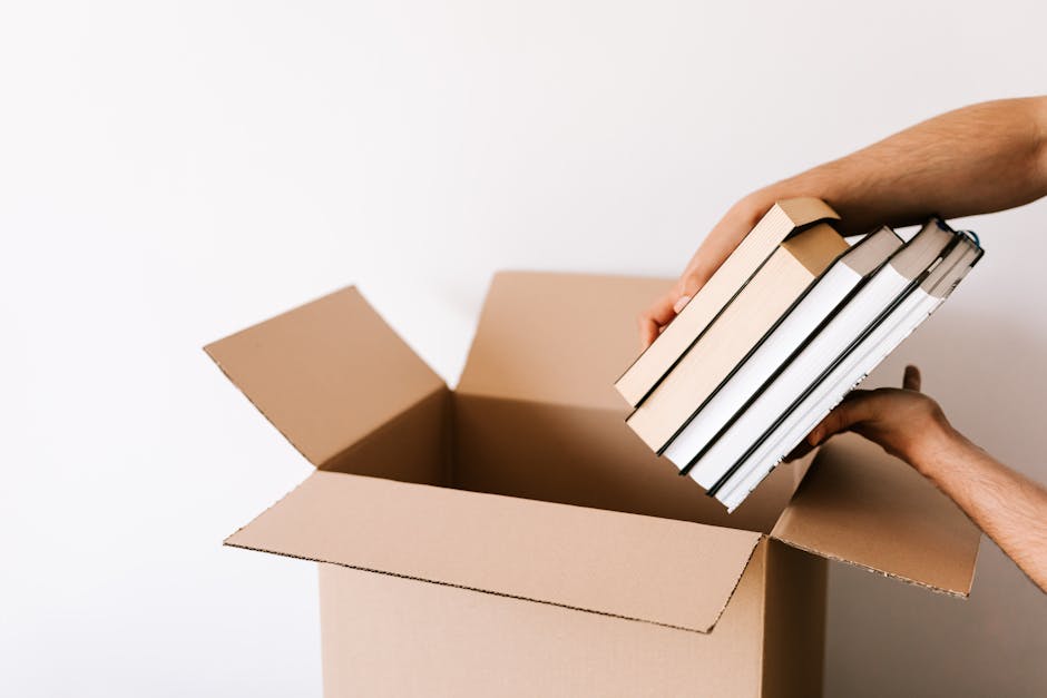 A person is placing a stack of six hardcover books into an open, light brown cardboard moving box. The person's hands are visible, one supporting the bottom of the books and the other steadying them as they are placed inside the box. The books are arranged vertically with the spines facing outward, featuring neutral and white covers with some metallic accents on the edges. The box is positioned on a flat surface against a plain white background, suggesting an indoor setting during a home relocation process. Nearby, a few other cardboard boxes are visible, indicating packing activity typical of furniture transport and packing and moving services. The image highlights careful handling of books and organized packing, reflecting professional removal and moving operations, such as those offered by Movers Brompton. Lighting is bright and even, emphasizing clarity and detail suitable for accessibility and SEO relevance.
