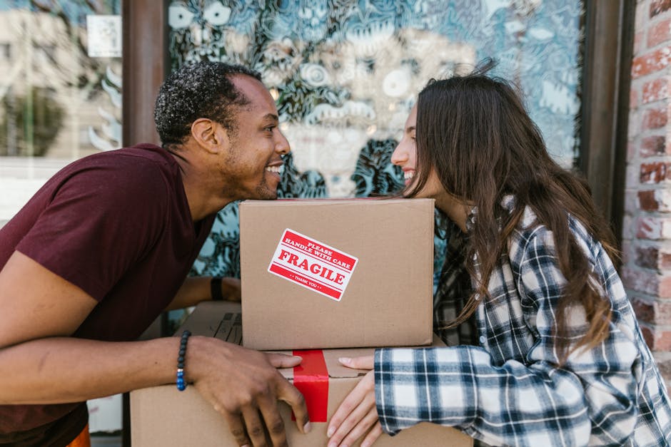 A man and woman outdoors, leaning towards each other with their foreheads touching, smiling during a house relocation or packing process. The man, wearing a maroon T-shirt and a beaded bracelet, is ho