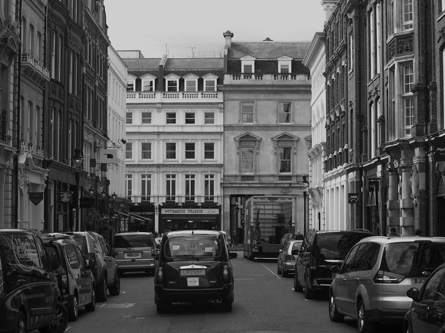A black and white photograph of a narrow urban street lined with multi-storey Victorian-style buildings, featuring ornate facades, large sash windows, and decorative architectural details. Several vehicles, including small vans, cars, and a delivery truck, are parked along the sides of the street on the pavement. In the foreground, a small black van is positioned centrally, with other vehicles parked on either side. Behind these, a loading process is visible, with a large cardboard box and packing materials situated on the pavement near a building entrance, indicating ongoing household or office furniture transport as part of a house removal or home relocation process. A trolley and moving blankets are also visible, supporting the loading activities. The scene depicts a typical street in Brompton, with natural light illuminating the surroundings, and the backdrop of residential buildings above the street level. This setting aligns with the services offered by Movers Brompton for removals, packing, and furniture transport within the Brompton Road area.
