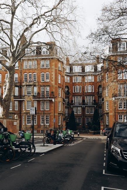 A view of a multi-storey red brick residential building on Brompton Road, with several large, leafless trees in the foreground and a parking area in front. The parking lot contains several green and black bicycles secured to bike racks, as well as a few parked cars, including a black vehicle on the right. The building features numerous white-framed windows, some open, and a series of small balconies. The street appears calm with minimal activity, and the scene is captured in daylight with overcast weather, highlighting the building's traditional architecture. This image illustrates the typical environment for house removals and packing services, with space for loading and unloading or moving furniture within the vicinity, supporting logistical planning for relocations through local streets highlighted on the Brompton Road guide on moversbrompton.co.uk.