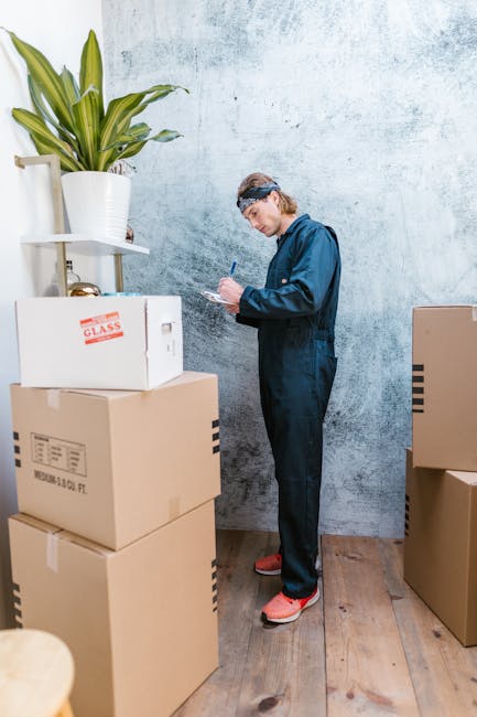 A person dressed in a dark blue work jumpsuit with a headband, standing indoors next to several stacked cardboard boxes, some labeled with fragile glass icons, in a room with a light textured wall. The individual appears to be taking notes or checking a checklist with a pen. To the left, there is a white shelf holding a potted plant with long green leaves. The floor is wooden, and additional boxes are visible to the right of the person, likely prepared for a home relocation or furniture transport. This scene depicts the packing and moving process, highlighting the importance of careful organization during house removals, as seen in professional moving services like those offered by Movers Brompton.