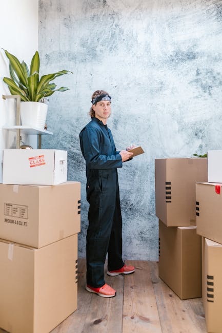 A man wearing a black cap and a white polo shirt is kneeling outdoors on a paved surface, carefully packing a cardboard box with writing tools and paperwork during a home relocation process. Surrounding him are several large cardboard boxes, some sealed and others open, containing various packing materials such as plastic wrapping, fabric covers, and smaller cartons. Behind him, a vehicle with an open rear door is visible, indicating loading or unloading activities typical of a furniture transport and moving service. The scene is well-lit with natural daylight, and the background includes a portion of the street and sidewalk. This image illustrates the packing and loading process involved in professional removals services, as provided by Movers Brompton, supporting effective house moves like those described on the Kings Road to South Kensington moving checklist.